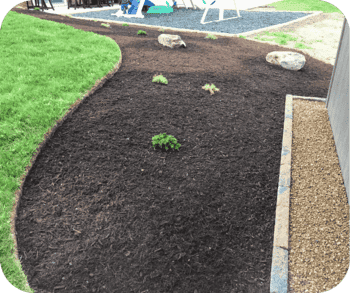 Freshly mulched garden bed with small plants and decorative rocks, bordered by a lawn and a gravel pathway near a play area.